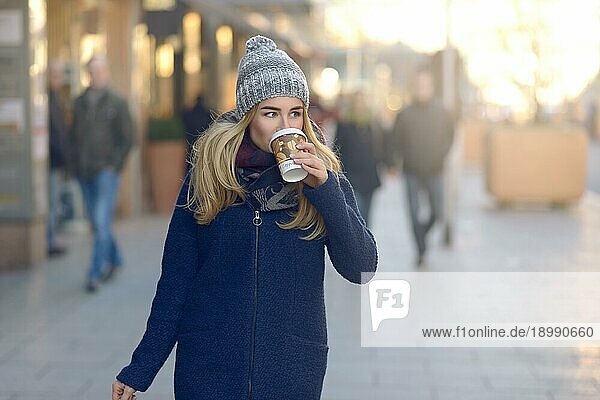 Blonde junge Frau mit Wollmütze  die Kaffee trinkt und auf dem Bürgersteig der Stadt spazieren geht  mit Sonnenstrahlen im Hintergrund
