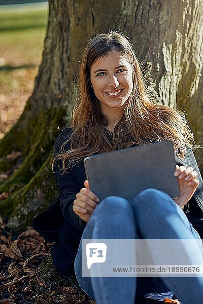 Glückliche attraktive junge Frau  die unter einem Baum sitzt  sich an den Stamm lehnt  ein Tablet auf den Knien hat und E Learning betreibt  und der Kamera ein freundliches  warmes Lächeln schenkt