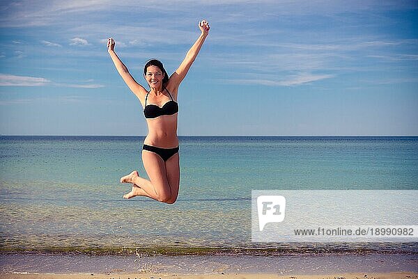 Agile attraktive junge Frau im Bikini hüpft vor Freude an einem schönen ruhigen tropischen Strand in der Sommersonne  während sie sich an der Natur und der Freiheit ihres Urlaubs erfreut