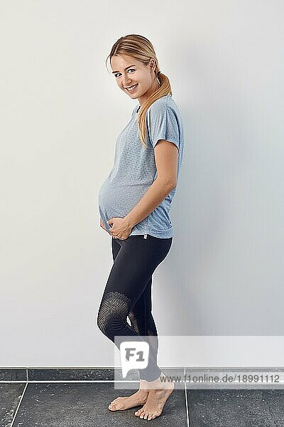 Seitenansicht einer fröhlichen  barfuß lebenden  schwangeren jungen Frau  die ihren Babybauch mit den Händen wiegt und sich mit einem warmen  glücklichen Lächeln vor einer weißen Wand zur Kamera dreht