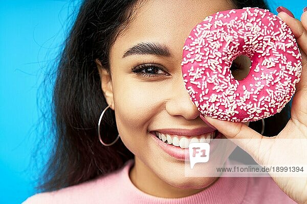 Nahaufnahme Porträt der schönen schwarzen Frau und Donut. Mädchen zeigt Donut vor ihren Augen auf blauem Hintergrund. Leckeres Essen  Diätkonzept