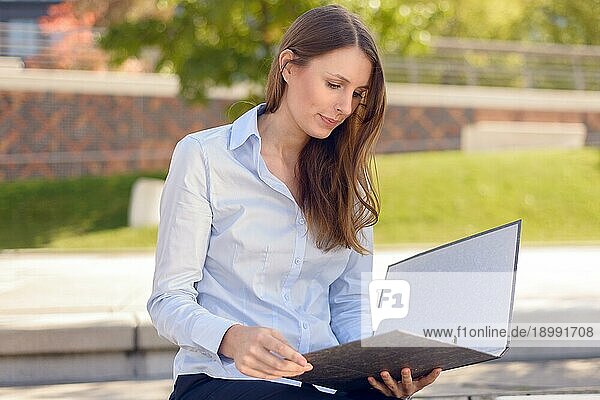 Attraktive Frau  die in einer Mittagspause oder auf einer Reise in eine fremde Stadt in einem Park auf einer Bank im Schatten eines Baumes eine Geschäftsakte liest oder ihre Notizen studiert
