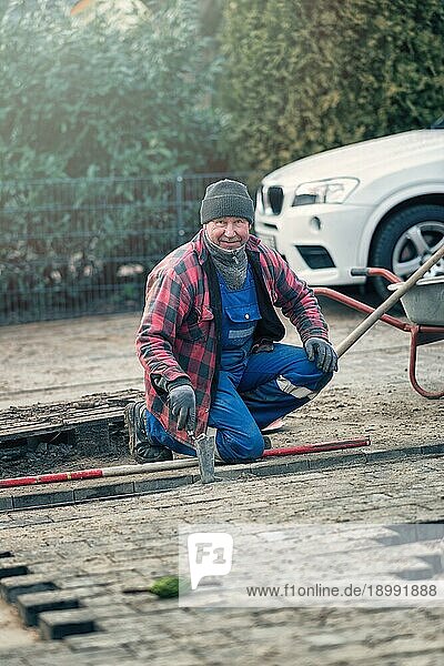 Fröhlicher Arbeiter in warmer Winterkleidung kniet am Straßenrand und verlegt Pflastersteine in seiner Latzhose und schaut mit einem freundlichen Lächeln in die Kamera