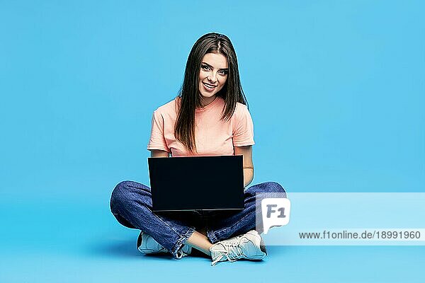 Porträt einer glücklichen jungen Frau  die mit gekreuzten Beinen auf dem Boden sitzt und an einem Laptop auf blauem Hintergrund arbeitet