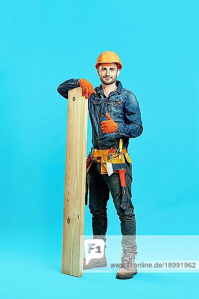 Ganzkörperporträt eines jungen  gut aussehenden Zimmermanns  der einen Stapel Holz hält und ein Daumen hoch Zeichen vor blauem Hintergrund zeigt und zur Kamera schaut