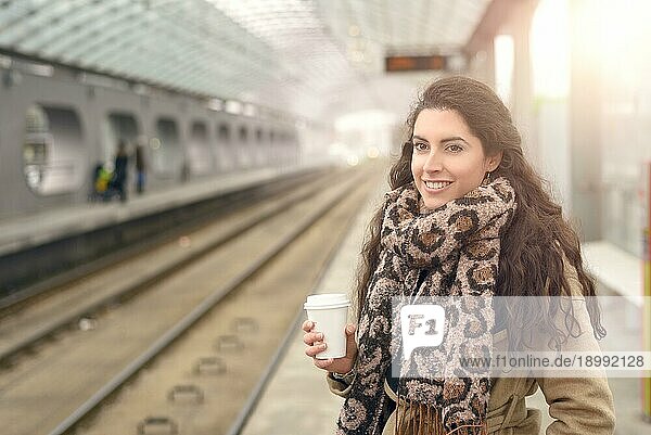 Einzelne lächelnde Frau in hellbraunem Wintermantel und Kaffeetasse in der Hand  während sie auf dem Bahnsteig einer S Bahn Station steht