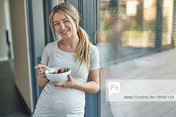 Glückliche  gesunde  schwangere  junge  blonde Frau  die an ein Terrassenfenster gelehnt steht und in die Kamera lächelt  während sie eine Schüssel mit frischem Obstsalat genießt