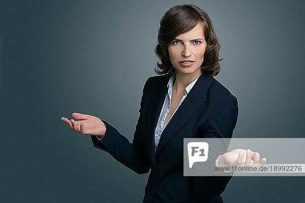 Halbkörperaufnahme einer verwirrten jungen Geschäftsfrau mit Händen in der Luft  die gegen eine graue Wand in die Ferne schaut