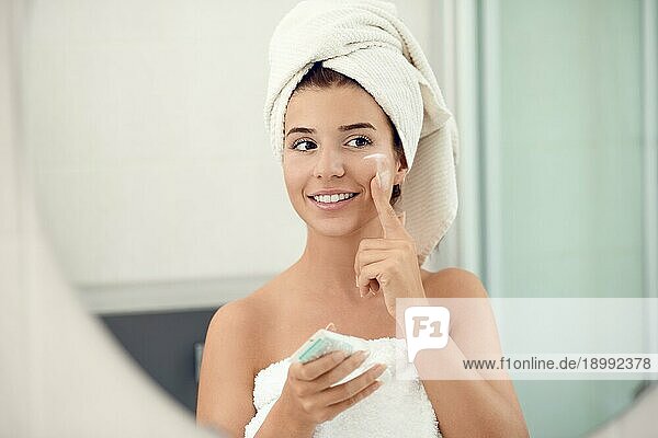 Hübsche junge Frau in weißen Handtüchern in einem Badezimmer  die der Kamera ein freundliches Lächeln schenkt  während sie ihre Wangen eincremt und ein Hautpflege und Hygienekonzept verfolgt