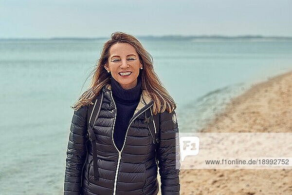 Attraktive Frau  die an einem bewölkten Tag am Strand spazieren geht  einen Rucksack und eine Jacke trägt und am Rande des Meeres innehält  um in die Kamera zu lächeln