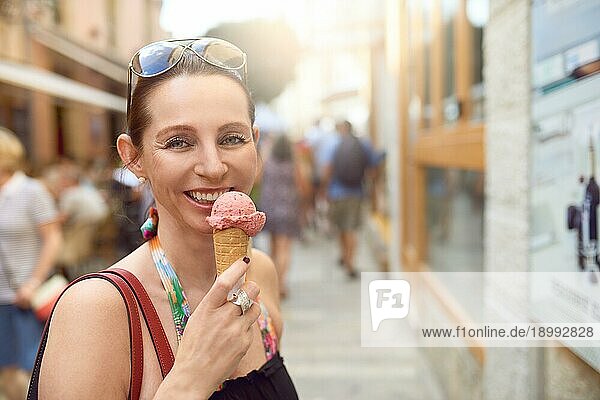 Lächelnde attraktive stilvolle Frau mit Sonnenbrille auf der Stirn  die ein Eis in einer Waffel im Freien in einer Fußgängerzone in einer Stadt genießt