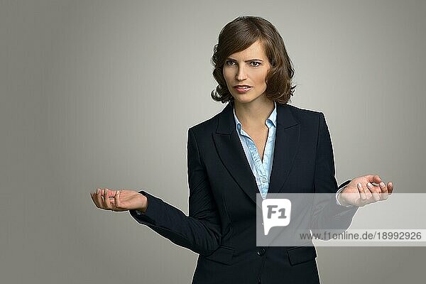 Halbkörperaufnahme einer verwirrten jungen Geschäftsfrau mit Händen in der Luft  die gegen eine graue Wand in die Ferne schaut