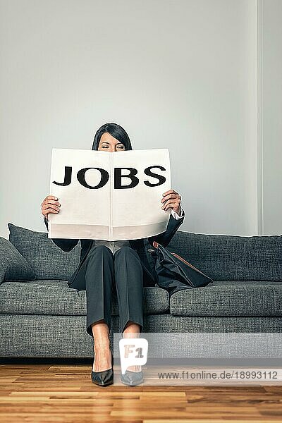 Stilvolle  berufstätige Frau  die auf einem grauen Sofa sitzt und die Stellenanzeigen in einer Zeitung liest  während sie aufgrund von Arbeitslosigkeit nach neuen Möglichkeiten  Veränderungen oder Arbeit sucht