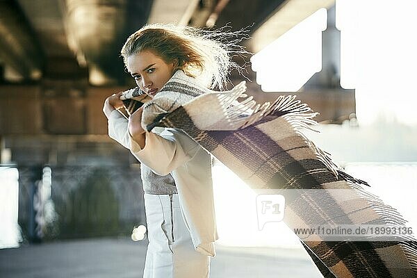 Schöne junge Frau mit Decke Blick zur Kamera auf windigen sonnigen Tag posiert auf städtischen Straße Hintergrund. Ausdruck  weibliche Schönheit  Vertrauen Konzept