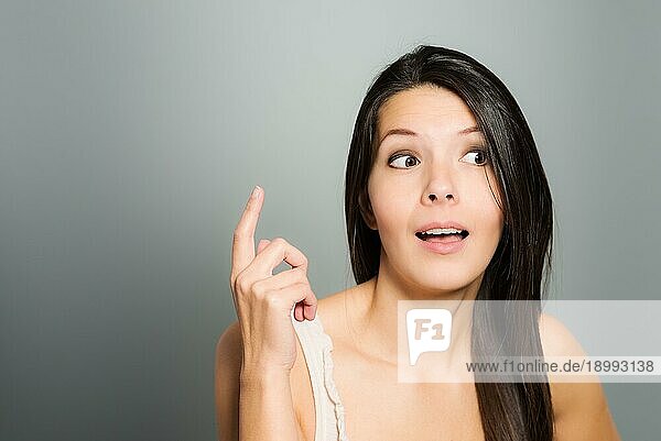 Attraktive junge Frau  die mit ihrem Zeigefinger signalisiert  daß sie entweder versucht  die Aufmerksamkeit auf sich zu lenken  oder ihren Finger zum Einlass erhebt  indem sie sich zu erkennen gibt  vor einem grauen Studiohintergrund