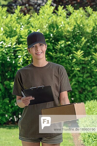 Ein lächelnder  attraktiver weiblicher Kurier mit einer Mütze  der ein Paket an die Tür eines Hauses bringt und ein Klemmbrett zur Unterschrift bereithält  um den Empfang zu bestätigen  vor einem Hintergrund aus frischen Gartenblättern