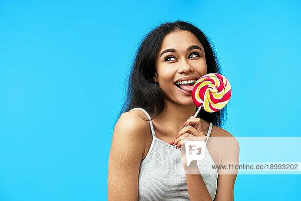 Glückliche junge schwarze Frau leckt bunten Lutscher auf blauem Hintergrund. Essen  Spaß Konzept