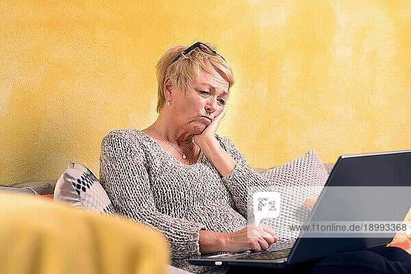 Attraktive Frau mittleren Alters  die zu Hause einen Laptop benutzt  während sie sich auf einem bequemen Sofa entspannt und nachdenklich auf den Bildschirm blickt  offener Blick