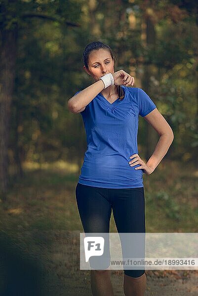 Eine Sportlerin wischt sich den Schweiß von der Stirn auf ihr Armband  als sie während ihrer Trainingsübungen auf einem Waldweg eine Pause einlegt