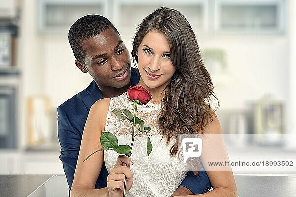 Hübscher schwarzer amerikanischer Freund  der seine hübsche Freundin mit einer Rosenblüte umarmt und in die Kamera lächelt  in der Küche stehend