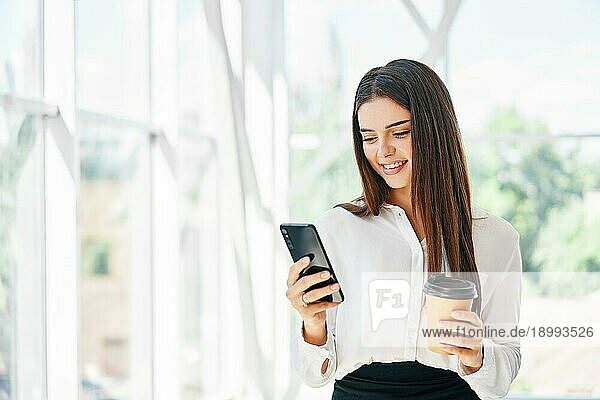 Hübsche junge Frau schaut auf Smartphone und lächelnd mit Tasse Kaffee in modernen kreativen Büro. Geschäftsfrau entspannen während der Pause Zeit