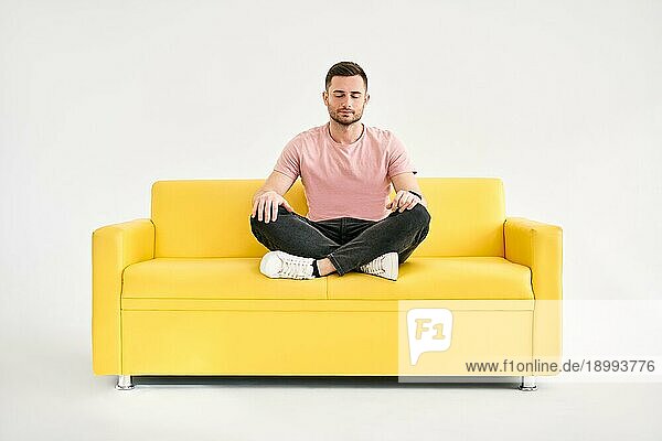 Hübscher junger Mann sitzt in Yogalotuspose und meditiert auf einem bequemen gelben Sofa vor weißem Hintergrund