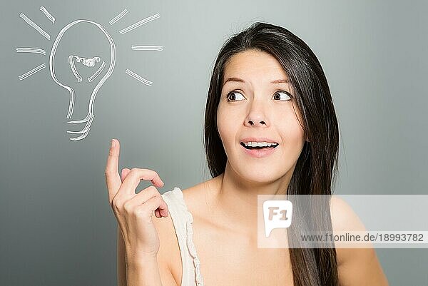 Attraktive junge Frau signalisiert mit ihrem Zeigefinger eine Idee in Form einer illustrierten Glühbirne  auf einem grauen Studiohintergrund