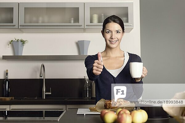 Glückliche attraktive junge Köchin  die den Daumen nach oben streckt  während sie in ihrer Küche steht und Kaffee trinkt  während sie einen frisch gebackenen Apfelkuchen vor sich hat