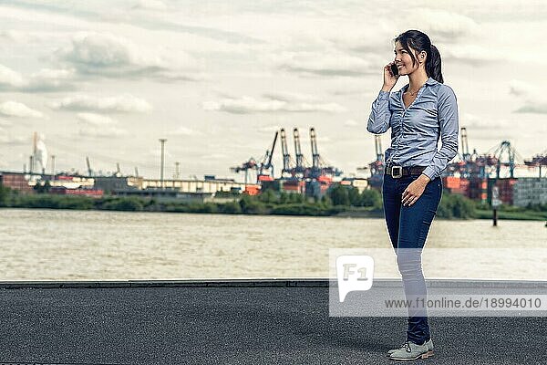 Attraktive brünette glückliche Frau trägt lässige Kleidung wie blaue Röhrenjeans und Hemd  während sie mit dem Handy spricht und am Ufer entlang des Flusses an einem bewölkten Tag geht