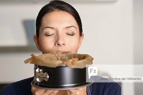 Attraktive Frau genießt den Duft eines frisch gebackenen Kuchens in einer Backform  die sie gerade aus dem Ofen genommen hat  wobei sie ihr unteres Gesicht mit geschlossenen Augen verdeckt