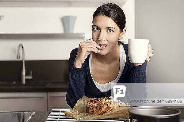 Junge Frau entspannt sich in der Küche mit einer Tasse Kaffee und probiert ihren frisch gebackenen Apfelkuchen  der auf dem Regal vor ihr abkühlt  während sie sich auf den Tresen lehnt und in die Kamera schaut
