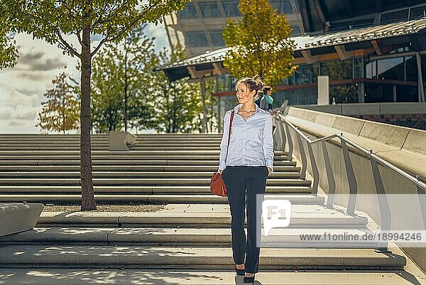 Schlanke  elegante junge Frau mit einer Handtasche  die eine Freitreppe aus Beton in einer Stadt mit modernen Geschäftsgebäuden im Hintergrund hinabsteigt
