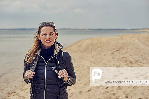 Attraktive  gesunde Frau  die an einem kalten  bewölkten Tag eine Strandwanderung genießt und lächelnd in die Kamera schaut  während sie die Riemen ihres Rucksacks hält