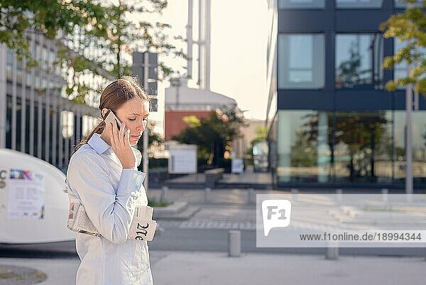 Junge Frau mit einer gefalteten Zeitung unter dem Arm  die in einer städtischen Straße steht und mit ernster Miene dem Gespräch am Handy lauscht  Seitenansicht