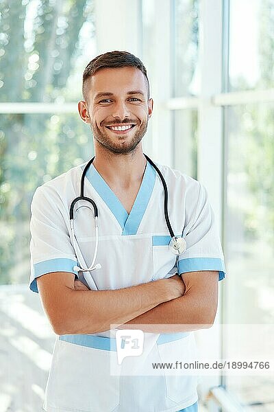 Porträt eines zuversichtlich lächelnden männlichen Arztes mit Stethoskop in medizinischer Uniform  die Arme verschränkt