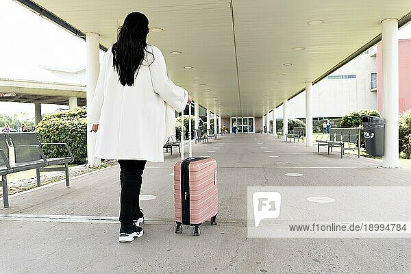 Rückenaufnahme einer Frau in Turnschuhen und Mantel  die zum Haupteingang eines Flughafens geht und ihren Rollkoffer rollt. Reisekonzept
