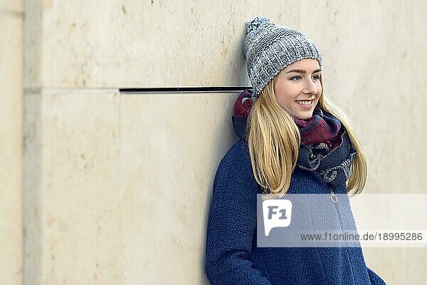 Lächelnde  attraktive  junge  blonde Frau in einem warmen  blaün Winteroutfit  die tagträumend mit dem Rücken an eine Außenwand gelehnt in die Ferne starrt