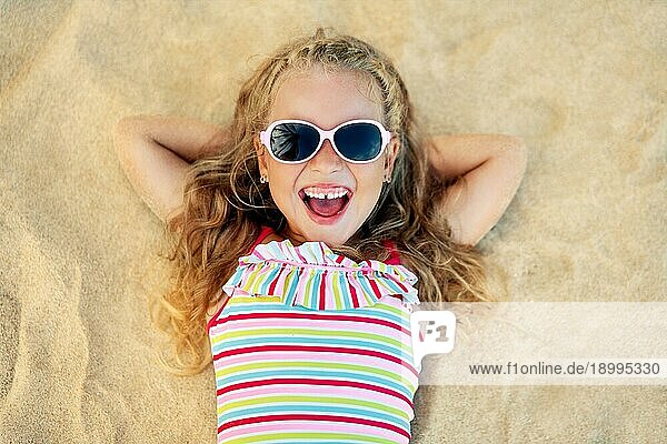 Draufsicht auf hübsches kleines blondes Mädchen mit Sonnenbrille am Sandstrand liegend während der Sommerferien. Meer entspannen  glückliche Kindheit Konzept