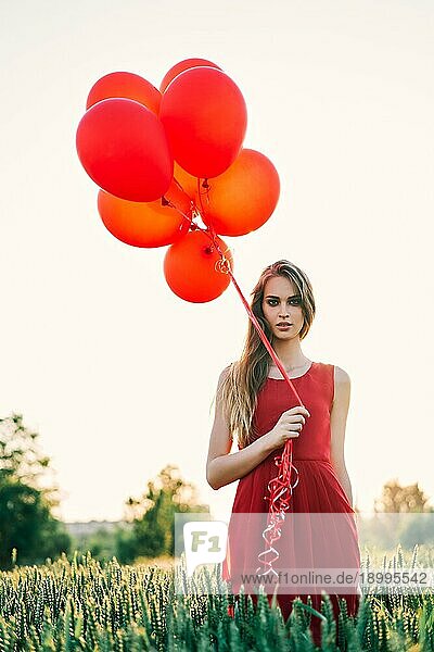 Junge schöne Frau in rotem Kleid posiert im grünen Feld mit roten Luftballons bei Sonnenuntergang. Freiheit  Spaß  Mode Konzept