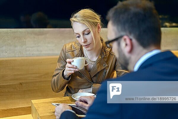 Geschäftsmann und Frau sitzen zusammen in einer Cafeteria und trinken Kaffee  wobei der Mann etwas auf seinem Tablet zeigt