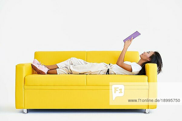 Hübsche asiatische Frau  die auf einer gemütlichen gelben Couch liegend ein Buch liest  Konzepte von Heim und Komfort  Hobby  Freizeit