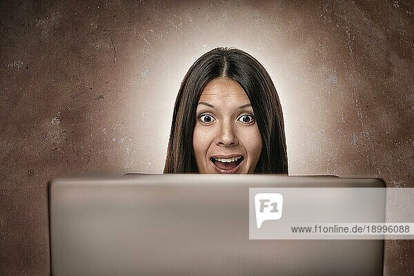Attraktive junge Geschäftsfrau blickt erstaunt und erfreut überrascht in die Kamera hinter ihrem Computerbildschirm  vor weißem Hintergrund
