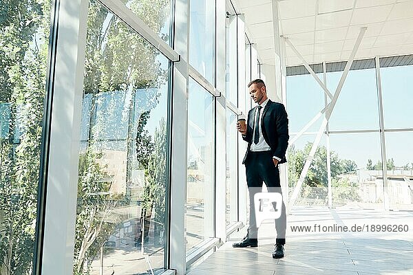 Full Länge Porträt der eleganten Geschäftsmann mit Tasse Kaffee in der Hand während der Pause in modernen kreativen Büro mit Kopie Raum. Entspannen  Erfolg Konzept