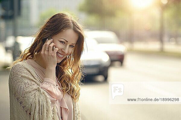 Attraktive junge Frau  die fröhlich lächelnd mit ihrem Mobiltelefon telefoniert  während sie am Rande einer städtischen Straße im frühen Morgenlicht mit Kopierraum steht