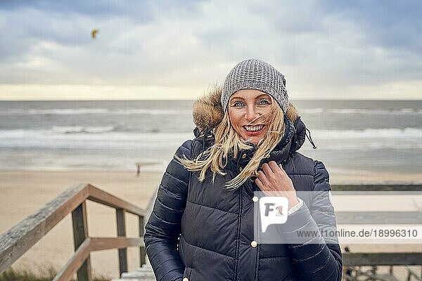 Frau mittleren Alters  die einem kalten Wintertag am Meer trotzt  steht auf einer Holzterrasse mit Blick auf den Strand an einem windigen Tag und lächelt glücklich in die Kamera
