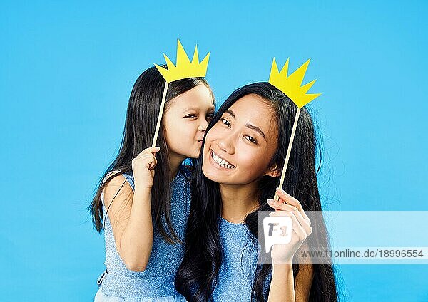 Nette kleine Tochter küsst ihre glückliche junge Mutter hält Papier Krone auf Stick auf blauem Hintergrund haben Spaß zusammen. Glücklich lustige Familie Konzept