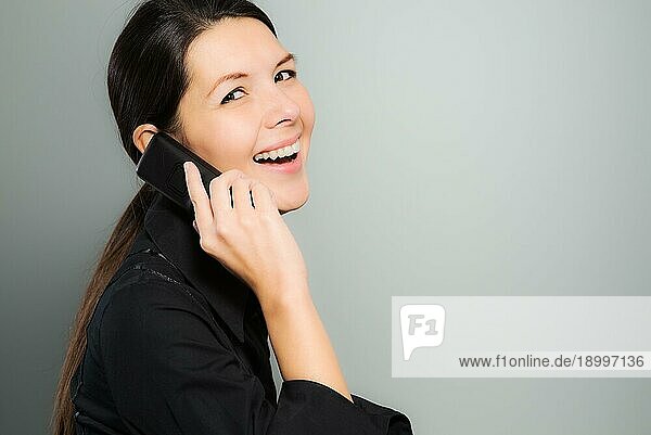 Attraktive junge Frau mit langen brünetten Haaren  die seitlich steht und vor Freude lacht  während sie mit ihrem Mobiltelefon auf einem grauen Hintergrund mit Leerzeichen plaudert