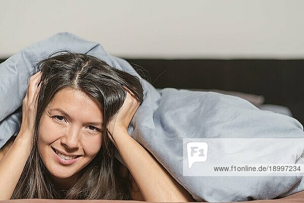 Lächelnde attraktive junge Frau  die einen faulen Tag genießt  auf dem Bauch liegend  unter einer warmen Bettdecke kuschelnd und mit einem glücklichen Lächeln in die Kamera blickend