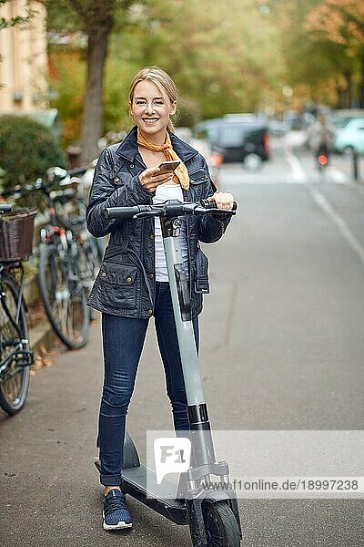 Junge Frau mit elektrischen Kick Scooter in der Straße der Stadt  Blick auf ihr Telefon und lächelnd. Öffentliche Verkehrsmittel Vermietung Service  moderne städtische ökologische Null Emissions Transport Konzept