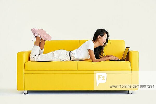 Hübsche lächelnde Frau mit Laptop auf einer gemütlichen gelben Couch auf weißem Hintergrund. Technologie  Freiberufler Konzept